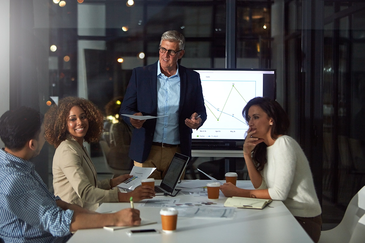 White man standing in front of a table full of smiling people in a conference room