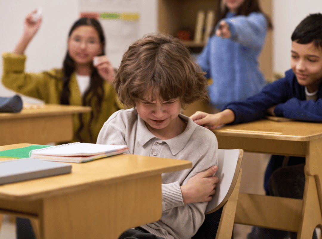 a young child sitting at a desk, looking down and upset, with other children around him