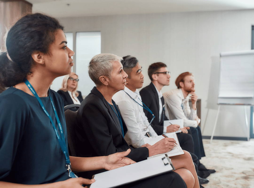 A row of professionals wearing lanyards sit attentively in a training room, some holding clipboards and notebooks, facing a presenter near a flip chart.