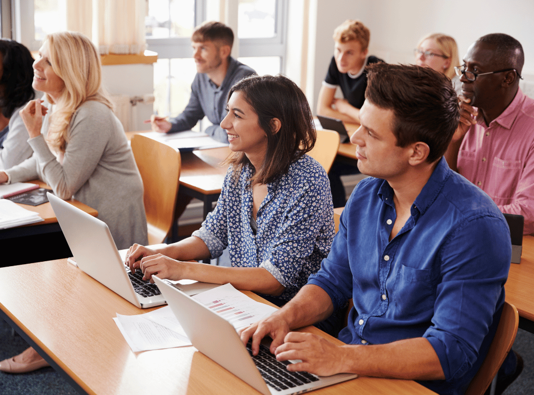 Adult learners sit at desks in a classroom, several typing on laptops with printed charts nearby, as they look toward the front of the room.