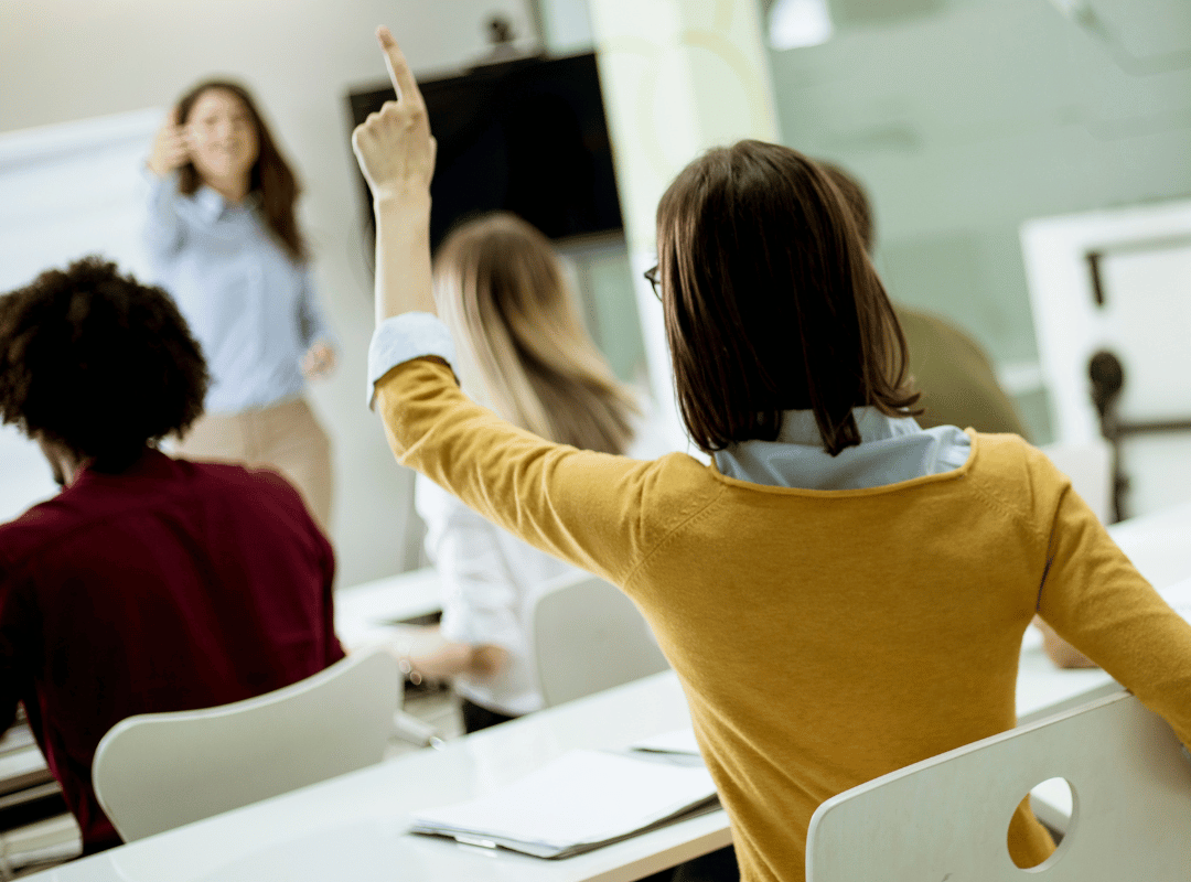 A participant viewed from behind raises her hand to ask a question while an instructor stands at the front of a training room.