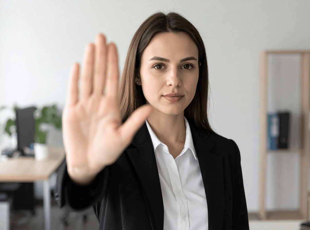 A woman in a dark blazer stands in an office and holds her palm out toward the camera in a calm stop gesture.